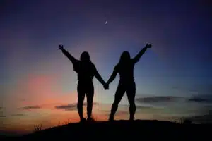 A stock photo of two people with their backs to the camera at dusk. We can see their silhouette against the sky, holding hands with their other hands raised.