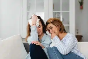 Two white women wearing light blue are sitting on a couch together crying.
