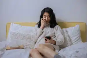 A young Asian woman sititng on a bed, looking at her phone and wiping tears off her face.