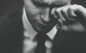A black and white close up photo of a man in a suit wiping tears away from his eyes.