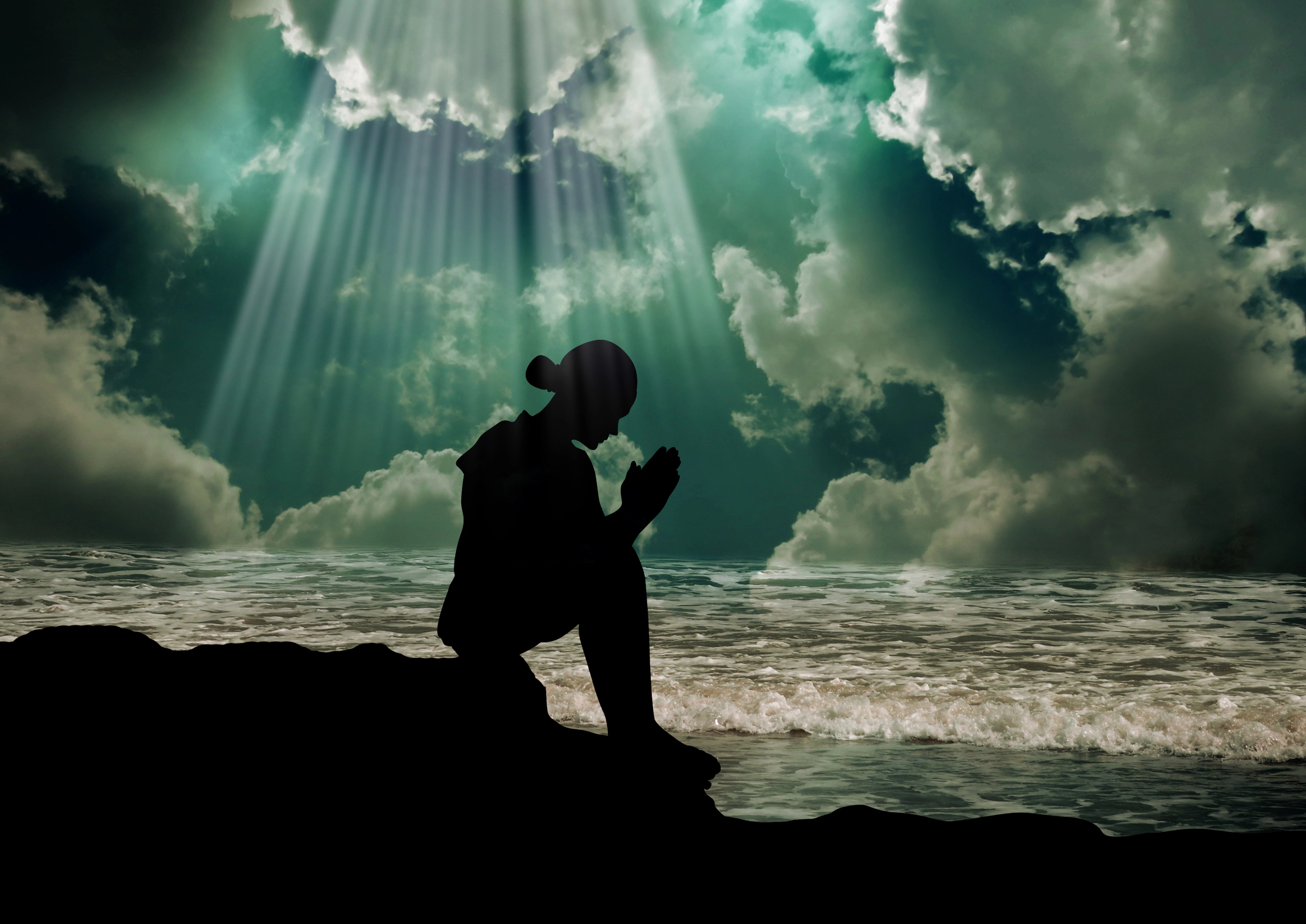 Silhouette of a person kneeling in prayer by the ocean, with rays of sunlight breaking through dramatic clouds in the sky.
