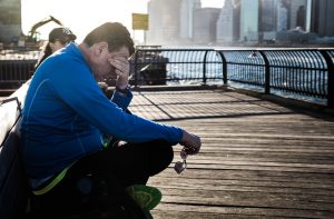 A white man wearing blue, sitting on a pier bench, rubbing his eyes with his hand