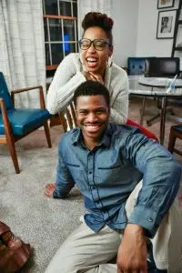 A stock photo showing a smiling Black couple sitting downk, with the man on the floor and the woman seated in a chair behind him. 