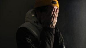 white man over a black background, wearing a yellow beanie and black jacket, and holding his hands over his face