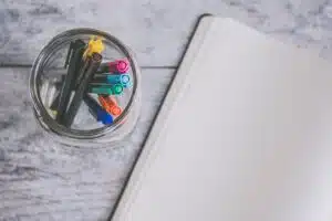 a cup of colorful markers next to an open blank page on a gray wooden background.