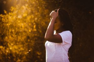 Black woman standing in front of a sunlit bush with her hand on her forehead, appearing distressed.