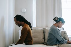 A stock photo of a Black couple sitting with their backs to one another on a made bed, looking distressed. 