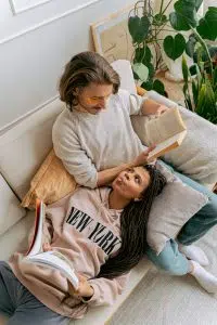 A stock photo of a couple sitting on a couch reading from above. The woman, who is Black, is laying across the couch, while the man, who is white, is sitting up. 