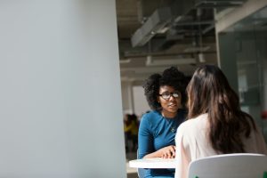 A Black woman sitting at a table in a workplace facing another woman whose back is to the camera. 