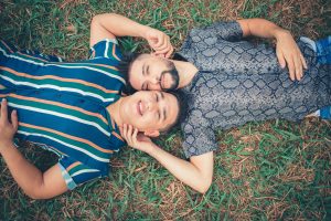 A gay couple laying on their backs in grass with their heads together, smiling.