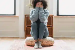 A stock photo of a Black woman sitting on a cushion on the floor, with both hands covering her face. | Why It’s Important to Talk About Grief (And 3 Tips for How to Do It) Blog