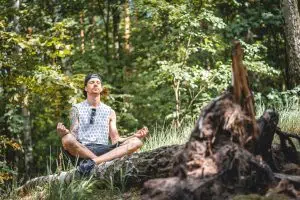 man wearing a backwards hat with sunglasses dangling from his white-and-blue tanktop meditating in the forest