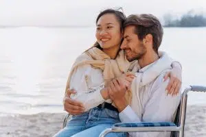 A stock photo of a couple on a beach - an Asian woman sits on the lap of a white man who is sitting in a wheelchair.