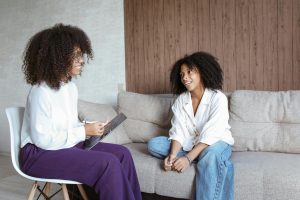 A Black psychologist providing psychological testing to a Black teen girl. The girl is sitting on a couch and the psychologist is sitting across from her. The psychologist is holding a clipboard on her lap. They are talking.