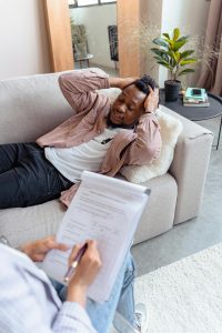 A Black man laying on a couch in a bright room, holding his hands on his head like he's distressed. A therapist is in the bottom left corner, holding a clipboard.