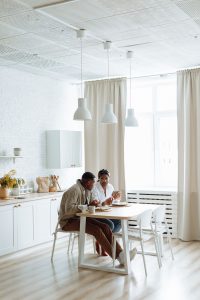 A photo of a Black couple in a bright sunny kitchen, sitting at a table eating breakfast food and talking to each other.