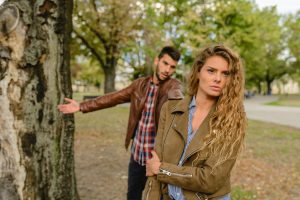 A white couple outside arguing, standing near a tree. The woman has her back to the man and is walking away, and they both look upset. 