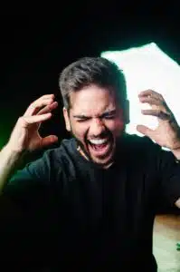 photo of a man in a dark room, yelling in anger. His hands are raised on either side of his face. 