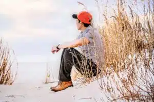 Image of a man in a red baseball cap sitting on a beach surrounded by sand and grass. He is looking away from the camera. 